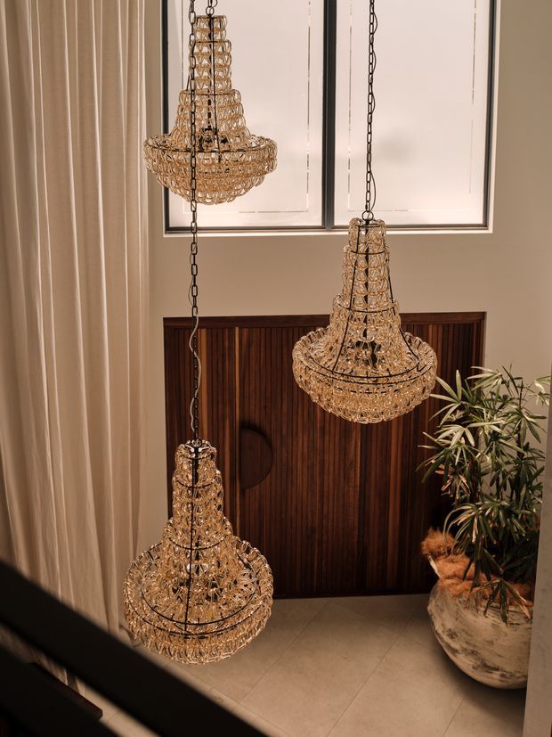 A warm, refined interior view featuring three ornate crystal tiered pendant chandeliers suspended on dark chains. They hang in front of framed wall artwork and a dark vertical timber-panelled feature wall, with full-height neutral curtains, pale tiled flooring, and a large indoor plant in a textured planter. The image appears to be taken from a stair or upper landing overlooking an entry or foyer.