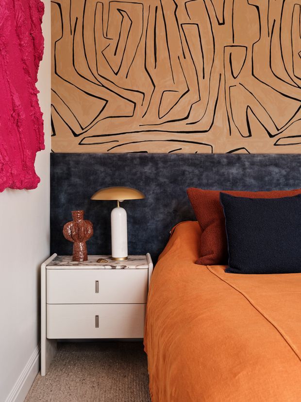 Close-up of a boldly styled bedroom corner featuring a white two-drawer bedside cabinet with a marble-patterned top, a sculptural white table lamp with a brass dome shade, and a small decorative vase. The bed is dressed in vivid orange bedding with rust and deep navy cushions. A dark charcoal textured wall panel and a large tan abstract artwork with black linework create a dramatic, eclectic composition, with a hot pink textured artwork partially visible at left.