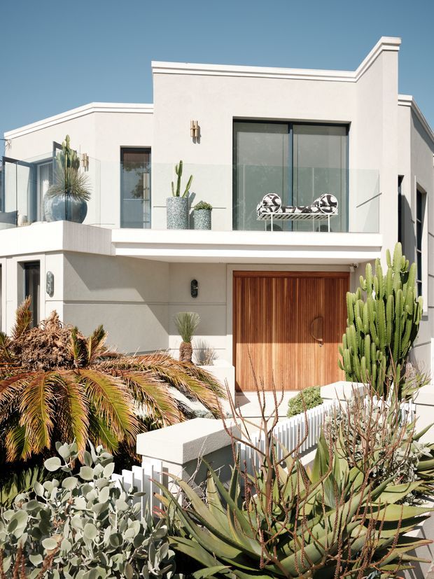 A bright contemporary two-storey residential facade with smooth white rendered walls, crisp parapet rooflines, dark-framed glazing, a large warm timber garage door, and a projecting upper balcony. The property is surrounded by dense drought-tolerant landscaping, including cacti, succulents and palms, with sculptural outdoor furniture visible on the balcony. The image is photographed from outside and is dominated by the building exterior.