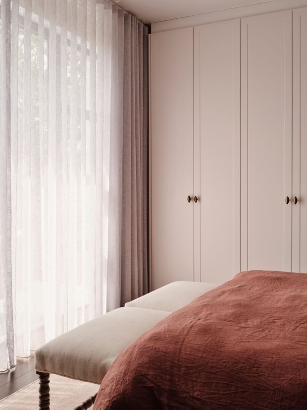 A softly lit bedroom corner with a large upholstered bed, blush-rose textured bedding, sheer full-height curtains layered with taupe drapes, and a wall of pale blush built-in wardrobes with small brass knobs. The room has a calm, refined, tonal palette and a contemporary fitted joinery design.