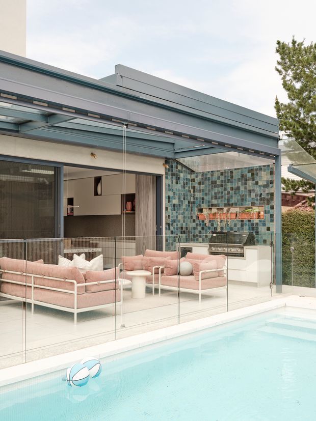 Contemporary poolside entertaining area with a glazed outdoor lounge and kitchen extension. The space features floor-to-ceiling glass walls, a slim grey metal roof and framing system, a turquoise mosaic tile feature wall, pale cabinetry, and soft pink outdoor sofas overlooking a bright swimming pool.