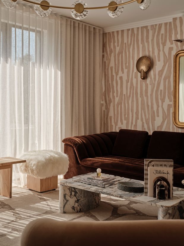 A richly styled living room with a deep brown velvet channel-tufted sofa, sculptural stone coffee table, plush white shearling ottoman, and warm timber side table. Sheer curtains filter abundant natural light, while patterned beige-and-taupe wallpaper, a brass sculptural wall light, ornate mirror, and statement ceiling fixture create a luxurious art deco-inspired atmosphere.