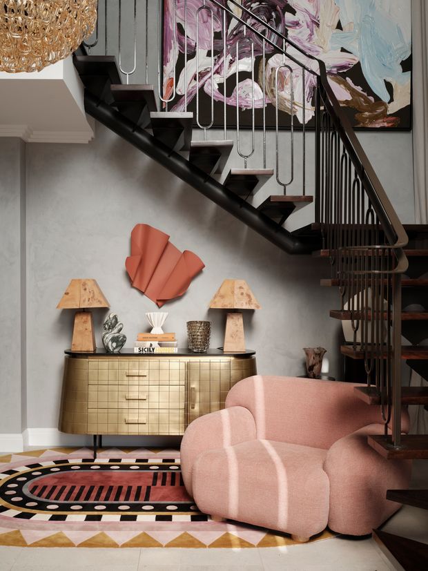 A richly styled contemporary interior featuring a sculptural black open-riser staircase with slender metal balustrades, a grey textured wall, large abstract artwork, and a brass-toned tiled sideboard. A curved blush-pink lounge chair sits over a bold geometric patterned rug, with sculptural lamps, decorative objects, and a statement chandelier adding an eclectic, artful character.