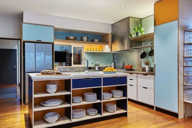 A warm, contemporary kitchen with a large central island featuring open shelving filled with crockery, integrated cabinetry in white, blue, timber and orange panels, stainless-steel appliances, and a stone-look benchtop. Timber flooring, pendant-free recessed lighting, a window, and plants complete the inviting space.