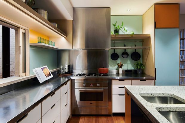 A compact, well-appointed contemporary kitchen with an L-shaped layout and central island. The space features dark stainless-steel appliances and splashback, pale blue-green walls, white and timber cabinetry, open shelving, integrated under-cabinet lighting, and a light stone island benchtop with an undermount sink. Pots and utensils hang above the cooktop, while a window and potted plants add natural character.