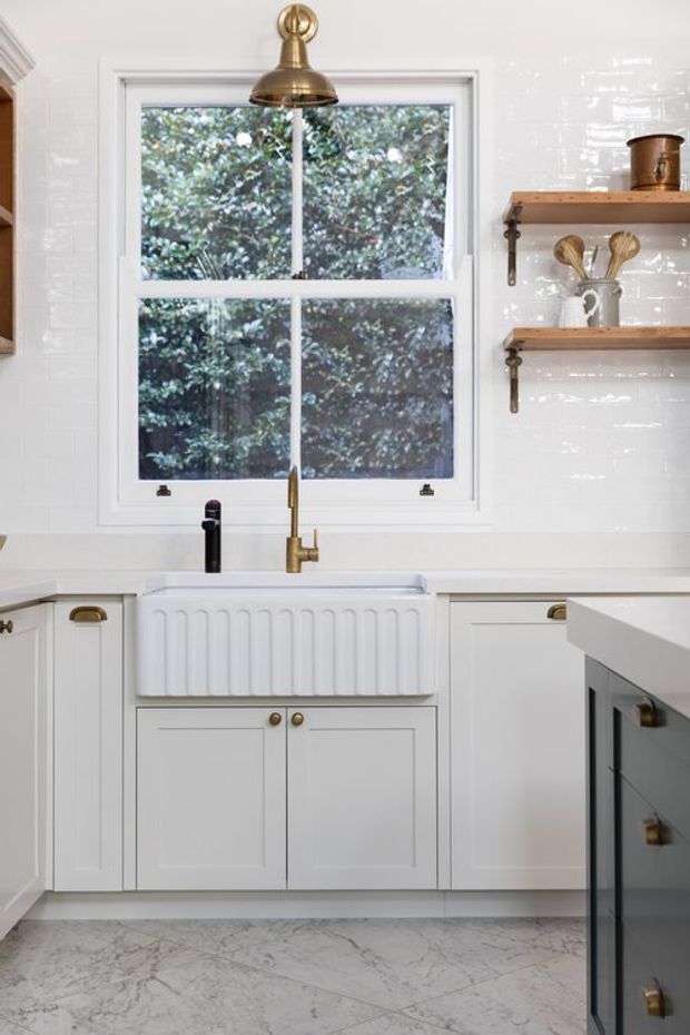 Bright, refined kitchen vignette centred on a white farmhouse-style apron-front sink beneath a divided-pane window. White cabinetry and glossy white tiled walls are paired with pale marble-look flooring and timber open shelves supported by dark brackets. Brass pendant lighting, tapware and hardware provide warm accents, while a dark island edge is visible in the foreground.