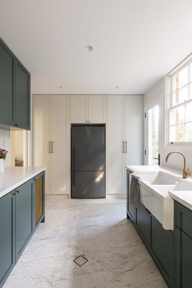 Bright, refined kitchen featuring deep blue-green lower cabinetry and a matching tall cabinet bank, warm white panelled cabinetry, pale veined stone flooring and benchtops, a large white farmhouse sink, and brushed brass tapware and handles. Full-height windows and a glazed exterior door bring in abundant natural light.