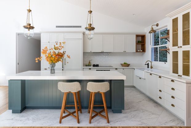 Bright, refined kitchen featuring a large white island with deep blue-green vertical-panel cabinetry, woven counter stools, white shaker-style cabinets, marble-look flooring and backsplash, brass hardware, pendant lights, and a built-in window-side cabinet wall.