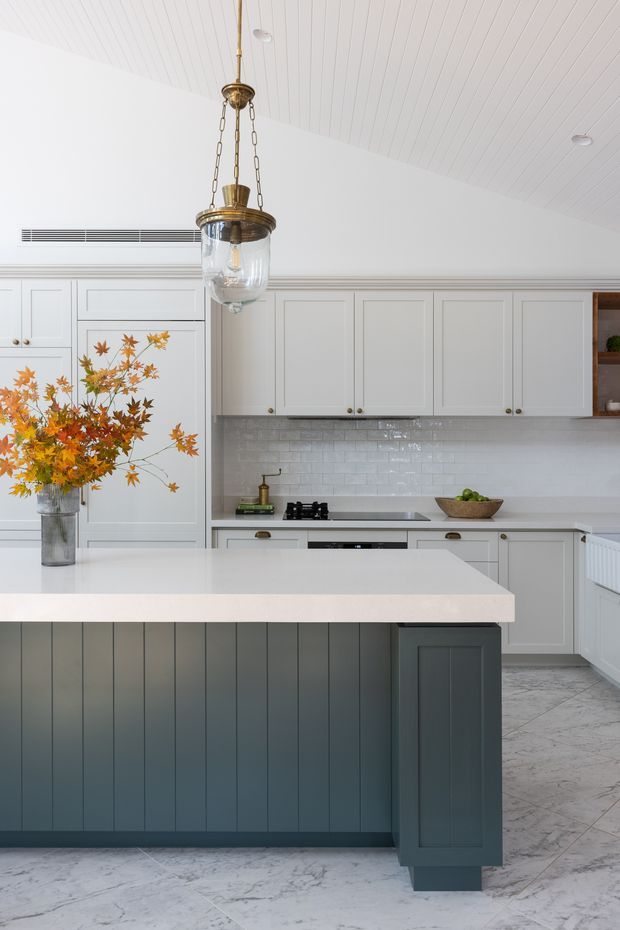 Bright, refined kitchen with white shaker-style cabinetry, a large dark blue-grey panelled island, pale stone benchtops, glossy white subway-tile splashback, and patterned pale stone flooring. A brass-and-glass pendant hangs above the island, while autumnal branches provide a warm decorative accent. The room has a high white panelled ceiling and abundant built-in storage.