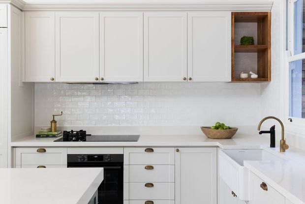 Bright, refined kitchen interior with white Shaker-style cabinetry, brushed brass hardware and tapware, a white subway-tile splashback, pale benchtops, integrated cooking appliances, and a timber open shelf niche. A large island occupies the foreground, with natural light entering through the window at right.