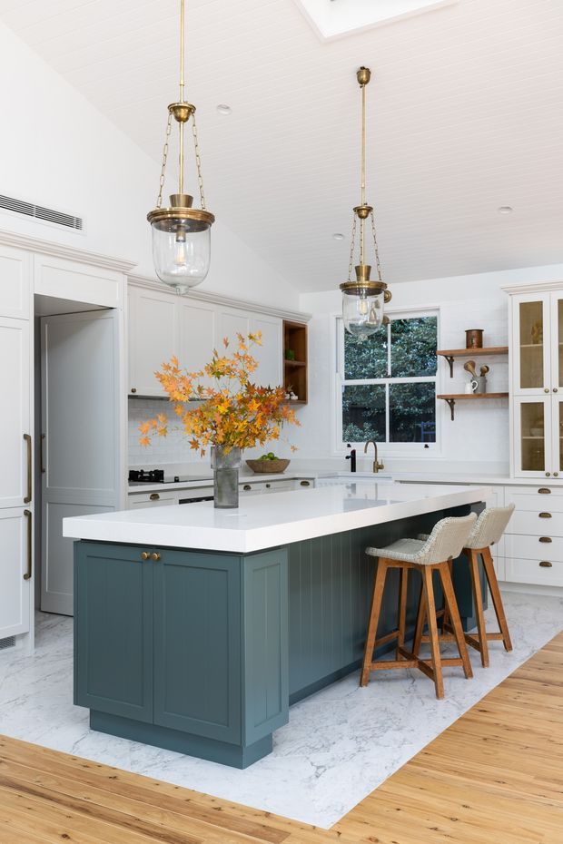 Bright, refined kitchen featuring a large deep teal-blue island with a white stone benchtop, white shaker-style cabinetry, marble-look flooring, timber flooring at the perimeter, brass-and-glass pendant lights, and woven timber bar stools. A window, open shelving, and tiled splashback add depth to the light-filled space.