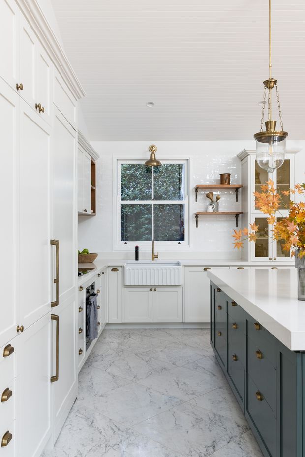 Bright, refined kitchen with extensive white Shaker-style cabinetry, a dark blue-grey island with a white stone benchtop, marble-look flooring, white tiled walls, brass hardware and pendant lighting. Open shelving, a sink beneath a window, and a floral arrangement add detail to the classic interior.