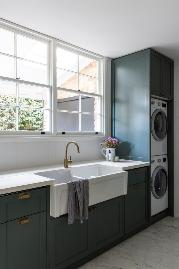 A bright, refined laundry room with large white-trimmed multi-pane windows, dark blue-green shaker cabinetry, white benchtops, a deep farmhouse-style apron-front sink, brass gooseneck tapware and hardware, and a stacked front-loading washer and dryer integrated into tall cabinetry. The room has white vertical wall panelling and pale veined stone-look floor tiles.