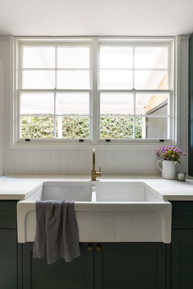 Bright, traditional-style kitchen sink area featuring a large white farmhouse apron-front double-bowl sink, dark green fixed cabinetry, a white countertop, white vertical wall panelling, brass-toned tapware, and large white-framed divided-light windows overlooking greenery.