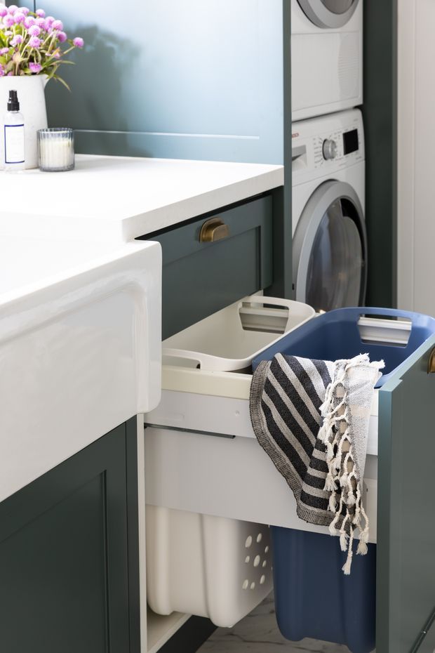A compact, thoughtfully designed laundry room with stacked front-loading washing and drying appliances, dark blue-green built-in cabinetry, white benchtops, pull-out laundry hampers, and baskets containing folded towels. A soft blue wall and small floral arrangement add colour and warmth.