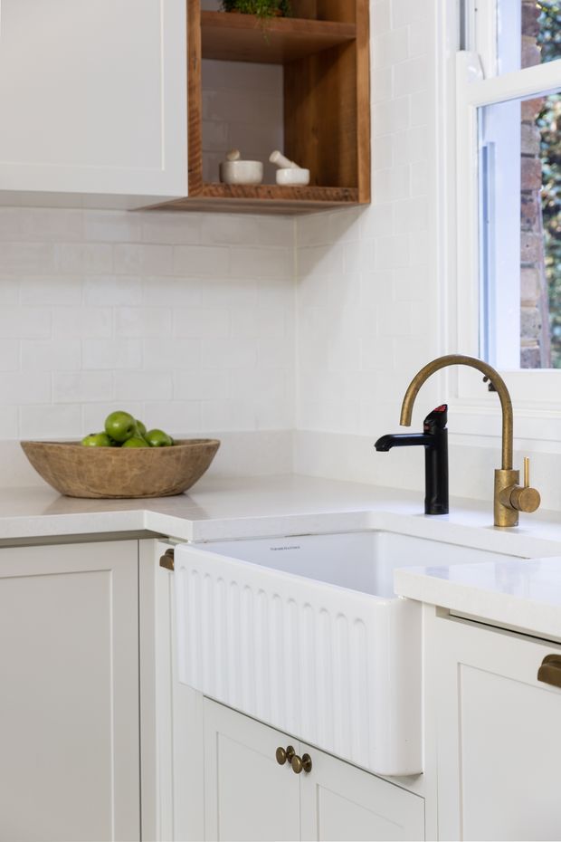 Bright, compact traditional kitchen corner featuring white shaker-style cabinetry, white subway tile splashback, white stone-look benchtops, a deep white farmhouse sink, and contrasting black and brass/gold tapware. A warm timber open shelf adds natural texture, while a white-framed window brings in daylight.