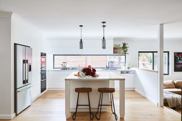 Bright, open-plan white kitchen with a central island and two stools, integrated appliances, pale timber flooring, pendant lights, and a partial view into an adjacent living area. Large windows provide abundant natural light, while white cabinetry and benchtops create a clean contemporary appearance.