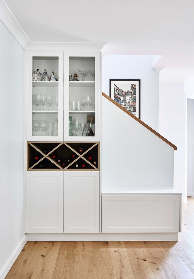 Bright, refined hallway and stair landing with a built-in white cabinetry unit incorporating glass-front display cupboards, wine storage, and lower paneled cabinets. A framed artwork sits beside a timber stair handrail, with pale engineered timber flooring and white decorative wall and ceiling trims.