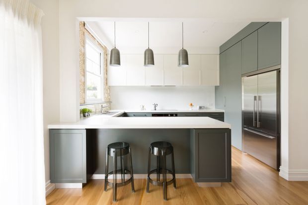 Bright contemporary kitchen with a large dark grey island and two bar stools, pale upper cabinetry, stainless-steel appliances, timber flooring, pendant lights, and a window with sheer curtains. The space has a clean, light-filled, refined appearance.