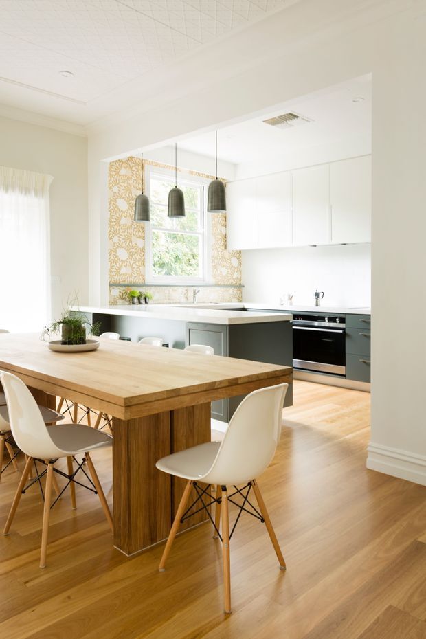 Bright open-plan kitchen and dining area with a pale timber dining table, white molded chairs, a central island, white cabinetry, stainless-steel appliances, timber flooring, patterned splashback wallpaper, and dark pendant lights. The room has a light, airy contemporary feel with subtle decorative detailing.