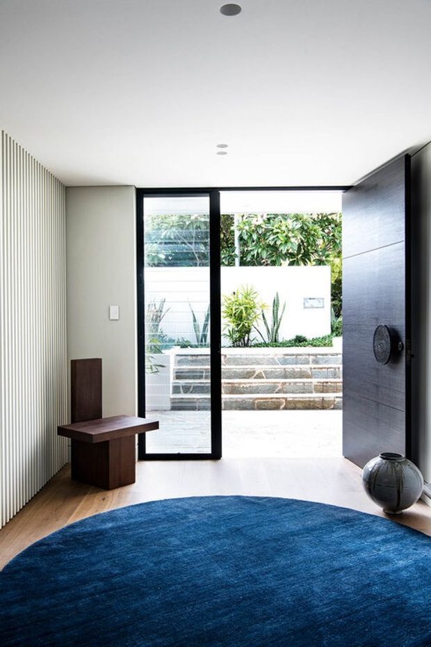 A bright, minimalist residential entryway with a large open black-framed glazed door leading to a landscaped courtyard. The interior features a white ceiling with recessed downlights, pale vertical wall panelling on one side, a dark timber feature door and wall on the other, warm timber flooring, a small dark timber console, and a large round deep-blue rug. Exterior stone steps, white rendered walls, and planting are visible beyond the doorway.