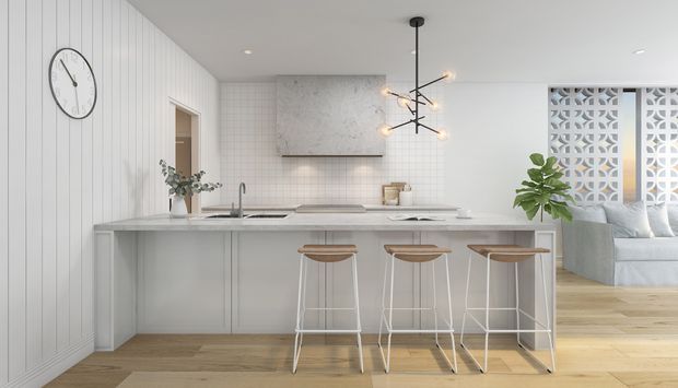 Bright open-plan kitchen with a long white island and three bar stools, pale timber flooring, white vertical wall panelling, tiled splashback, integrated cabinetry, and a sculptural black-and-brass pendant light. A living area with a sofa is visible beyond a decorative breeze-block screen.