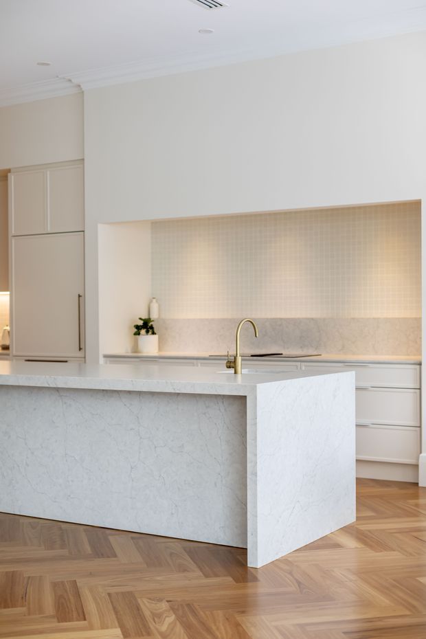 Bright, refined kitchen featuring a large waterfall island clad in pale veined stone, soft beige built-in cabinetry, a brass-toned gooseneck tap, and a subtle small-format tiled splashback with integrated lighting. The room has a white coffered-style ceiling with decorative moulding and a warm timber parquet floor laid in a herringbone pattern.