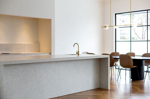 Bright open-plan kitchen and dining area with a large pale stone island, integrated cooktop and sink, softly lit tiled splashback niche, timber parquet flooring, and a dark-framed gridded window. A linear brass pendant light hangs above the dining table with brown upholstered chairs.