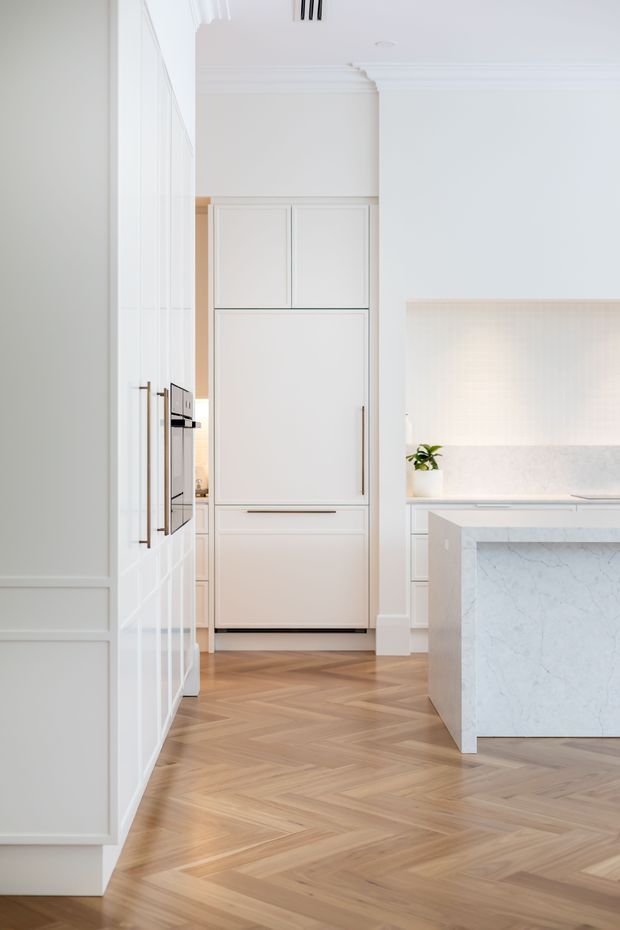 Bright, refined kitchen interior viewed from an adjoining passage, featuring white panelled cabinetry, integrated appliances, a pale marble or stone island and backsplash, decorative wall mouldings, and warm timber herringbone flooring. The space has a clean, light-filled classic-contemporary appearance.