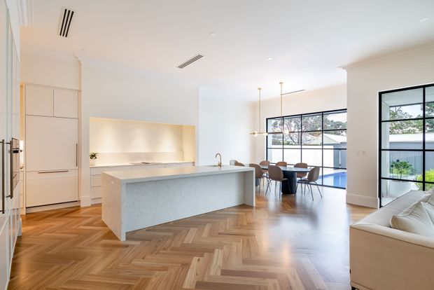 Bright, open-plan contemporary kitchen and dining area with a large pale stone island, integrated white cabinetry, herringbone timber flooring, pendant lights, and expansive black-framed glazing opening to an outdoor pool area. A partial living room is visible at right.