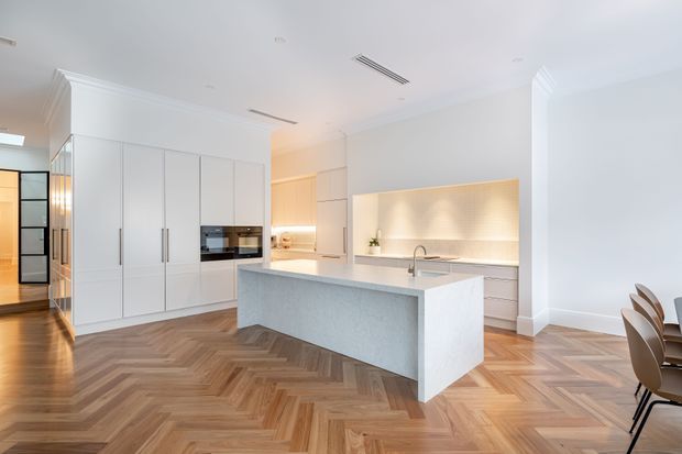Bright contemporary kitchen with a large pale stone island, integrated white cabinetry, built-in appliances, illuminated open shelving, and a timber herringbone floor. The open-plan space also includes a dining area and a refined neutral palette with decorative ceiling cornices.