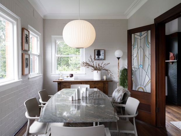 Bright, refined dining room with a long marble dining table surrounded by modern upholstered chairs. White-painted brick walls, timber-framed windows, decorative wall art, a large spherical pendant light, and a dark timber doorway with stained-glass panels create a blend of contemporary furnishings and traditional architectural detailing.