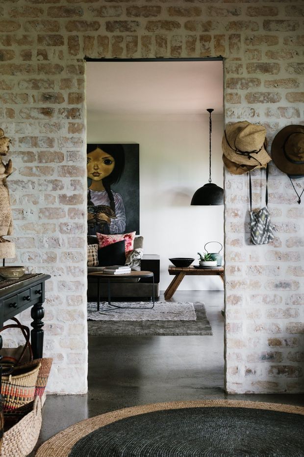 A view through a wide doorway framed by exposed pale brick walls into a relaxed living room. The room contains a dark sofa with colourful cushions, a low timber coffee table, a black dome pendant light, layered rugs, indoor plants and artwork. Hats and a patterned bag hang on the brick wall, while part of a dark timber console and woven baskets are visible in the foreground. The palette is warm, earthy and eclectic with an industrial-rustic character.