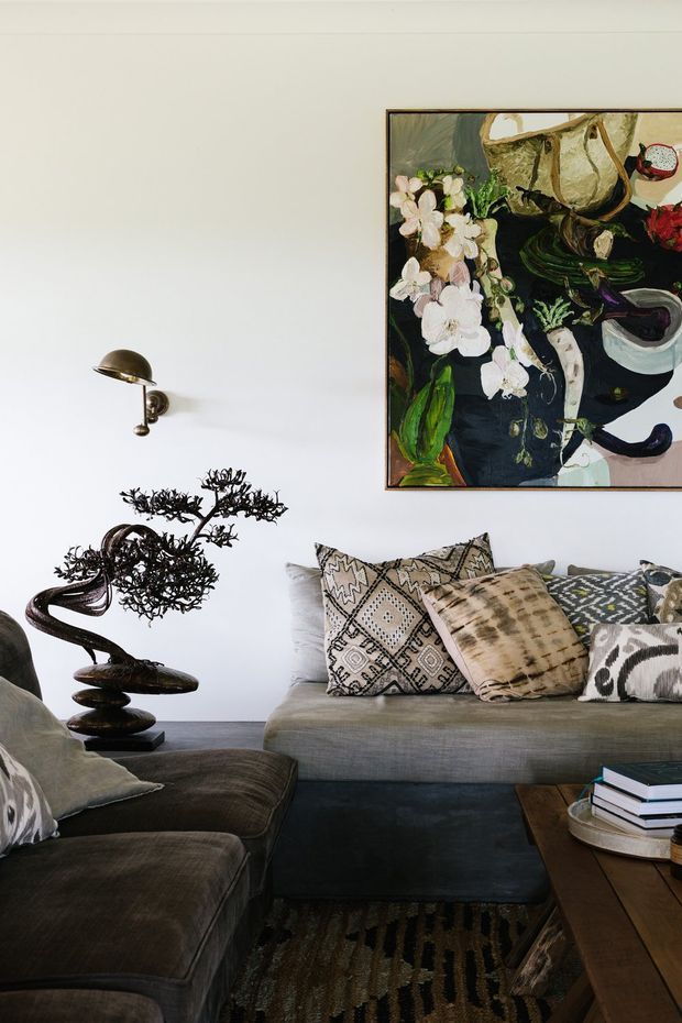 A refined living room with a grey sectional sofa and matching armchair, layered patterned cushions, a dark coffee table, textured rug, sculptural bonsai-like ornament, wall sconce, and a large expressive floral painting. The space has a neutral, moody palette with eclectic decorative elements and strong contemporary styling.