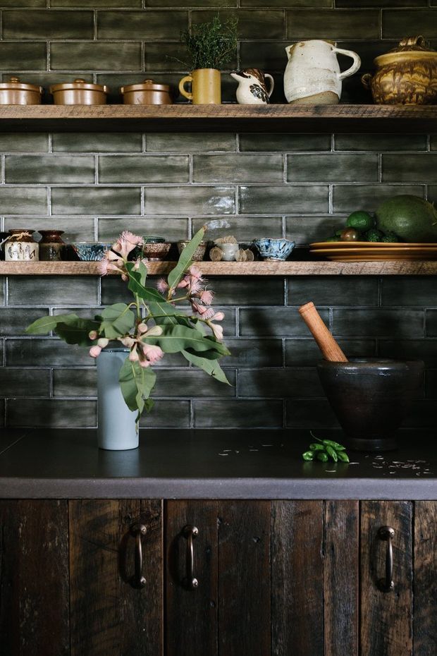 A close-up still-life view of a rustic kitchen or pantry-style wall with dark green-grey glazed rectangular brick tiles, two timber floating shelves, a dark stone or composite benchtop, and heavily textured dark timber cabinetry with black metal handles. Copper pots, ceramic vessels, a vase of leafy pink flowers, fruit, and a mortar and pestle add an artisanal, moody character.