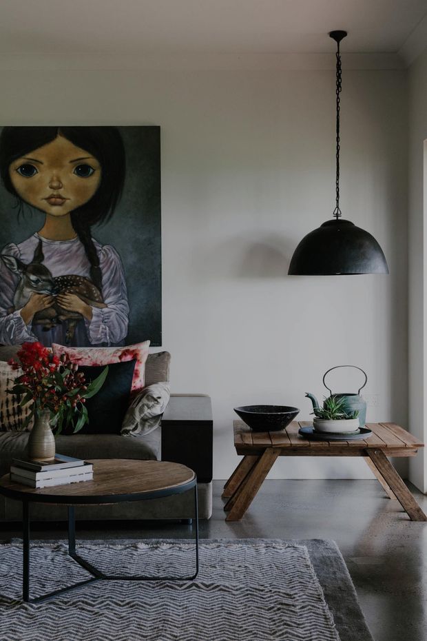 A moody, styled living room with a grey upholstered sofa, patterned cushions, a round timber coffee table, a rustic trestle-style side table, indoor plants, and a large whimsical portrait artwork. A black dome pendant light hangs over the room, with polished concrete flooring and a textured monochrome rug.
