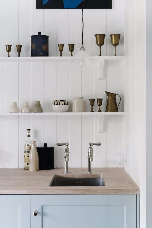 A compact butler's pantry or bar area with white vertical wall panelling, open display shelves, brass and ceramic vessels, a timber benchtop, polished metal bridge-style taps, and a small undermount sink. Pale blue shaker cabinetry sits below the counter, creating a restrained traditional coastal feel.
