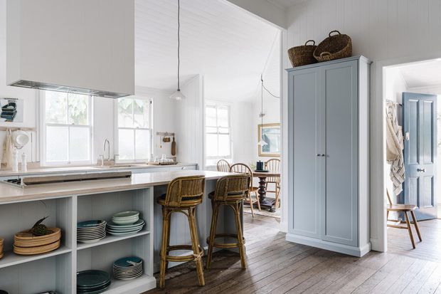 Bright, open-plan kitchen with a pale grey island, white cabinetry and walls, open shelving displaying dishes, woven counter stools, and a tall built-in storage cupboard. Timber floors extend into an adjoining dining area, while a blue-painted door and baskets add character. The space has abundant natural light and a relaxed coastal-country feel.