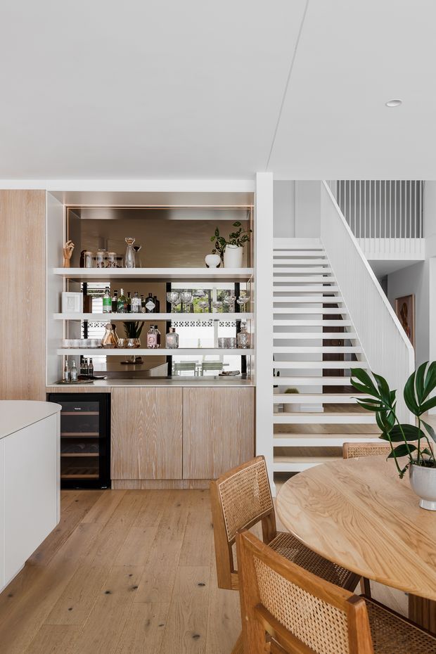 Bright contemporary open-plan dining and bar area with a warm timber floor, built-in timber cabinetry, open shelving stocked with glassware and bottles, and a pale stone or light benchtop. A round timber dining table with woven cane chairs occupies the foreground, while an open white staircase with vertical balustrade detailing rises beside the joinery. The room has a clean, airy palette of white, light wood and warm neutral tones.
