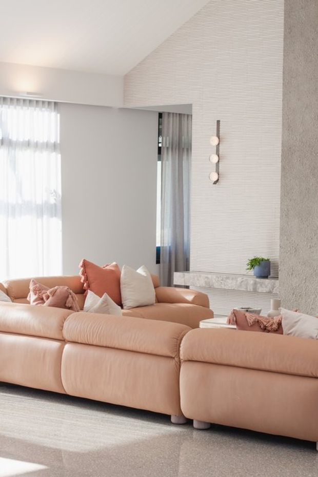 Bright contemporary living room featuring a large blush-pink sectional sofa, soft neutral cushions, sheer floor-to-ceiling curtains, and a textured white feature wall with a linear fireplace and minimalist pendant lights. The room has a refined, warm, softly layered aesthetic with abundant natural light.