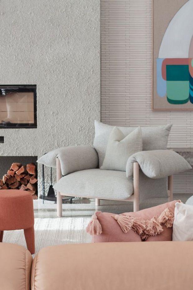 A softly styled living room with a pale upholstered armchair, blush and peach seating, a textured light wall, a linear tiled fireplace surround with stacked firewood, and a colorful abstract artwork. The space has a warm, refined, contemporary feel with layered neutral and pastel furnishings.