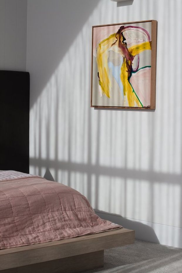 A softly lit bedroom corner with a low timber bed, dark upholstered headboard, muted dusty-rose quilt, grey carpet, and white vertical-panel walls. Strong sunlight filtered through blinds creates dramatic linear shadows across the wall and floor, while a colourful abstract artwork provides a focal point above the bed.