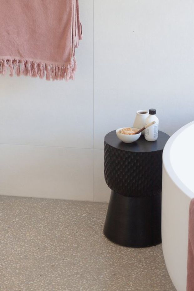 A softly styled contemporary bathroom corner featuring a freestanding white bathtub, pale wall tiling, terrazzo-look flooring, and a black textured cylindrical side table holding toiletries and a bowl. A dusty pink fringed towel adds warmth and colour.