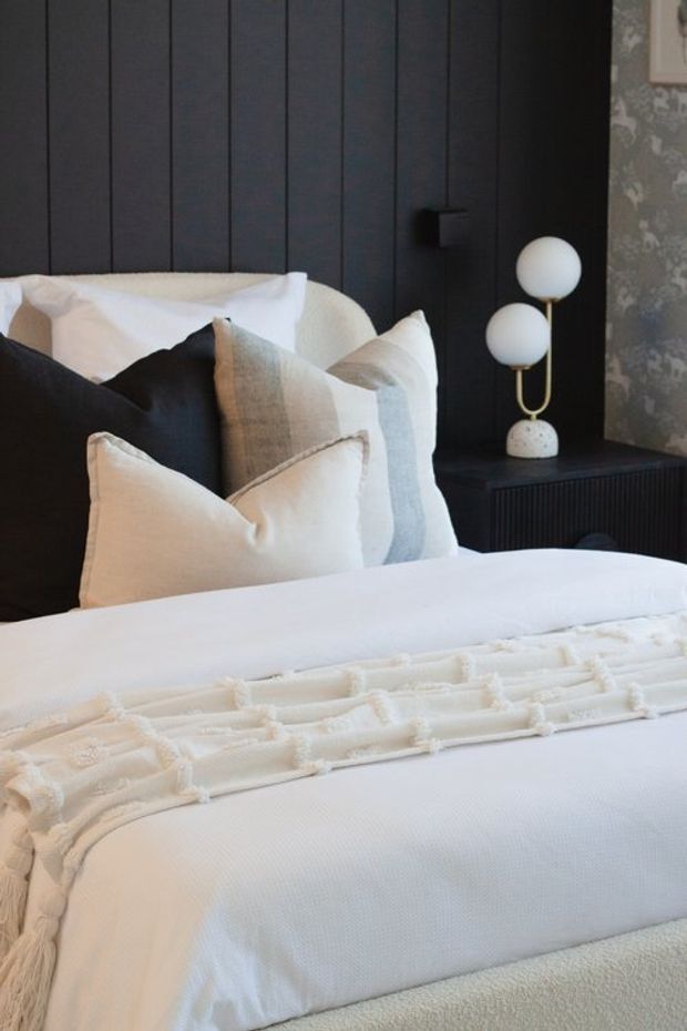 A close-up bedroom interior featuring a neatly made bed with layered white, cream, grey, and black pillows and textured white bedding. The bed is set against a dark vertically panelled feature wall, with a black ribbed bedside cabinet and a sculptural double-globe table lamp. A patterned grey wallpaper panel is visible at the right edge.