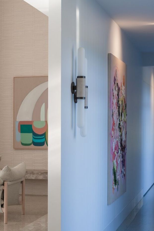 A refined interior hallway with softly blue-lit, vertically textured walls, a sculptural wall sconce, and colourful abstract artwork. An adjacent living area is partially visible through the opening, showing neutral upholstered seating, textured wall treatment, and a marble or stone fireplace surround. The corridor has a polished, gallery-like atmosphere with a cool, muted palette and colourful art accents.