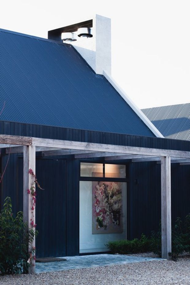 Exterior view of a dark blue contemporary building with a steep corrugated metal gable roof, white chimney, and a weathered timber-framed covered entry or verandah. The entrance has glazed doors and windows, with gravel paving and restrained planting around the facade.