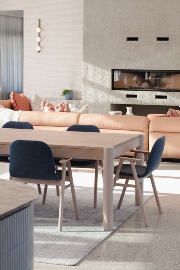 A bright, contemporary open-plan living and dining space. A pale timber dining table with dark blue upholstered chairs sits on a light rug in the foreground, while a soft peach sectional sofa faces a textured light wall with a long built-in fireplace. Neutral ribbed wall detailing, floor-to-ceiling curtains, sculptural pendant lighting, and strong daylight create a refined, warm interior.