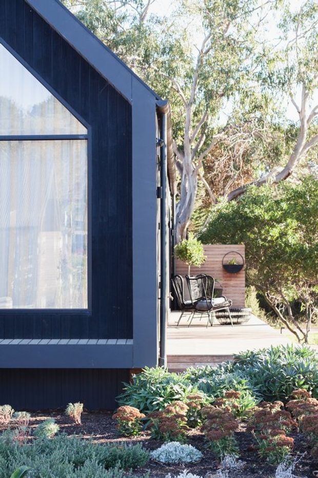Exterior view of a compact dark-clad house with a steep gabled roofline and large glazed opening, beside a timber deck furnished with outdoor chairs. Layered low planting and mature trees surround the building, creating a private garden setting.