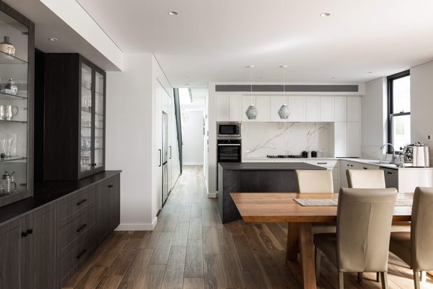 A bright, contemporary open-plan kitchen and dining area with white full-height cabinetry, a dark island, marble-look splashback, integrated appliances, timber flooring, and a substantial timber dining table. Dark built-in display cabinetry lines the left wall, while large windows bring in natural light.