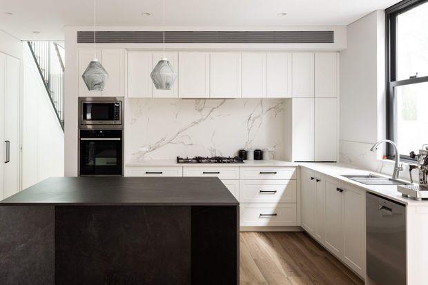 Bright contemporary kitchen with white full-height cabinetry, integrated black appliances, a large dark island, marble-look splashback, timber flooring, pendant lights and a stainless-steel sink area beside a large window. A stairway is partially visible at the left.